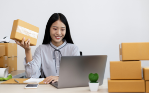 A person holding a brown cardboard box sits at a desk with a laptop, surrounded by stacked packages and office supplies.