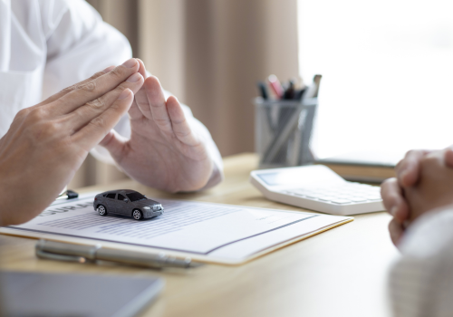 A person gestures while discussing a car-related contract, with a small toy car placed on the document in front of them.
