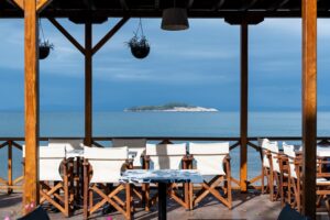 A serene view from a wooden patio featuring empty tables and chairs overlooking a calm sea and a distant island under a cloudy sky.