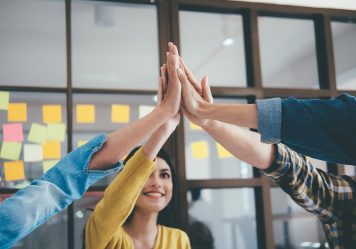 A group of hands reaching toward each other in celebration, with colorful sticky notes visible in the background.